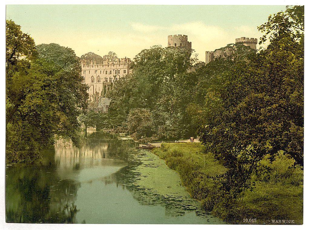 The image depicts a picturesque landscape featuring an old castle with prominent towers amidst lush greenery. The castle appears to be constructed of stone, exhibiting medieval architectural elements such as battlements and crenellations on its upper sections. It is set back from the water's edge where a calm river reflects parts of the structure and surrounding foliage. Verdant trees frame both sides of the scene, with one tree partially obscuring the castle on the left side.

The photograph has a vintage appearance, suggesting it may be an early 20th-century postcard or print based on its yellowish tint and aged quality. The text at the bottom right indicates "Warwick," likely referencing Warwick Castle in England's West Midlands region. This landmark is known for its historical significance dating back to Roman times when a fortress was established there.

The overall composition of this image exudes tranquility, capturing an idyllic moment that highlights natural beauty alongside human-made heritage.