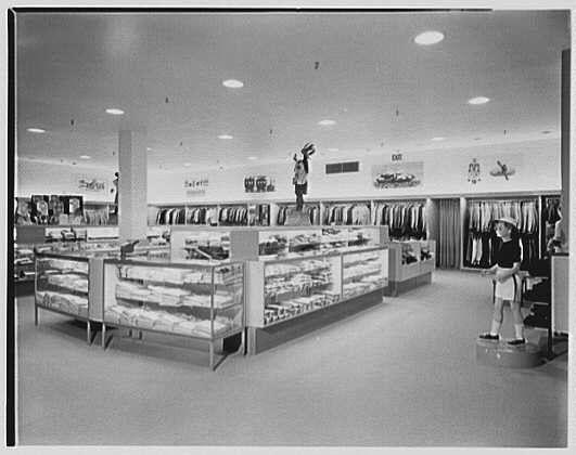 The image depicts an interior view of a clothing store, likely from the mid-20th century. The layout includes various sections with different merchandise categories, such as suits and dresses displayed on shelving units or in glass cases. A few individuals are visible: one person is reaching up to grab something off a higher shelf while another stands at an information desk near the entrance.

The store's design features include recessed lighting fixtures mounted on the ceiling, creating a well-lit environment. Promotional banners hang above some of the shelving units and glass cases, possibly advertising specific items or sales events. The monochrome tone suggests that this photograph was taken with black-and-white film during its time period.

This image is related to "Lord & Taylor," indicating it may have been captured at one of their business locations in West Hartford, Connecticut on May 25th, 1953 by Gottscho-Schleisner, Inc. It serves as an illustrative visual representation for the historical context and atmosphere of a mid-century clothing store environment.

Further information about this image can be found at: <https://images.loener.nl/gottscho-schleisner/full/691f/691f00fb450e57a2675039be.jpg>.