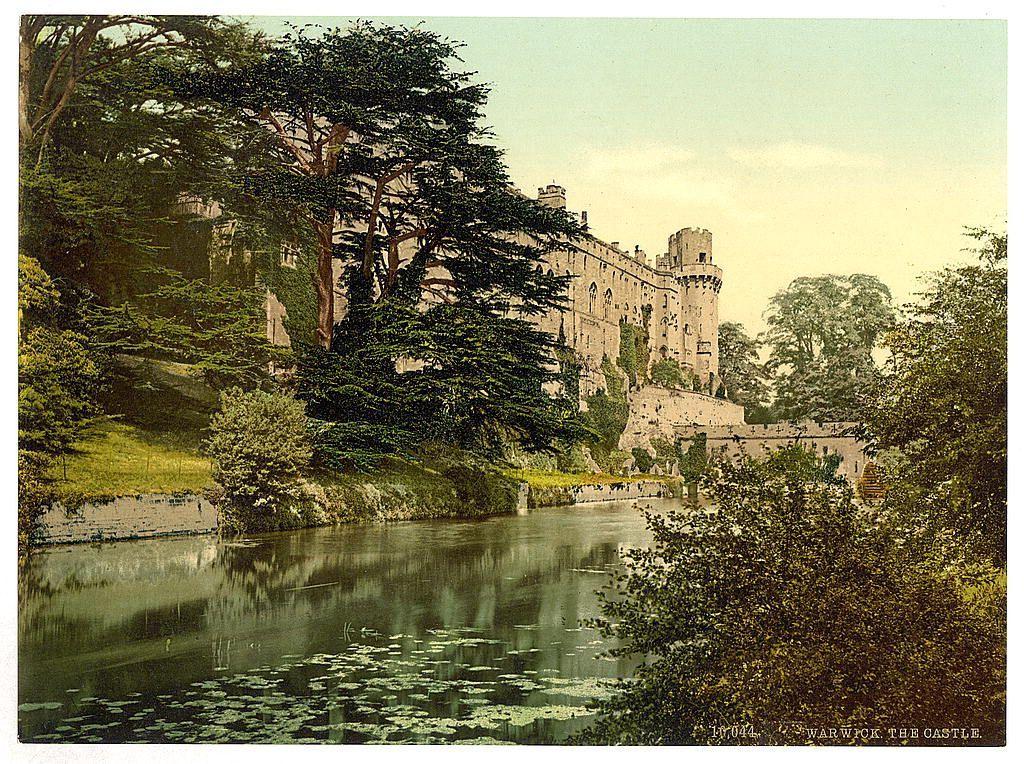 The image depicts a historic castle situated beside the River Avon in Warwick, England. The photograph is aged and has a sepia tone typical of early 20th-century printmaking processes known as photochroms. It features an impressive medieval stone structure with fortified towers and battlements along its walls. Surrounding trees frame the building, suggesting that it may be situated on elevated ground or within a park-like setting. The river is calm, reflecting parts of the castle's façade and vegetation from surrounding greenery. In terms of historical context, Warwick Castle has been occupied since Roman times; however, much of its current structure dates to medieval periods when such castles served as both residences for nobility and fortifications against invaders. This particular image likely originates around 1890-1900 based on the caption "Warwick The Castle."