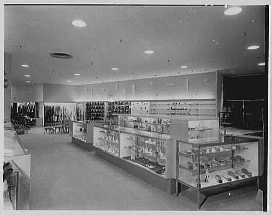 This black and white photograph depicts an interior view of a store, specifically focusing on the men's shop area. The image appears to be from around 1953, as indicated by the date in the description "May 25." The setting is likely associated with Lord & Taylor, a business located in West Hartford, Connecticut.
The photograph showcases various shelves and display cases containing merchandise such as neckties, belts, cufflinks, sunglasses, shoes, and other accessories. These items are neatly arranged on open racks or within glass-fronted cabinets for customers to browse through. The lighting fixtures can be seen mounted on the ceiling, casting a well-lit environment throughout the store.
In addition to this main area of focus, there is another section visible in the background that appears to have more products displayed, possibly related to women's fashion based on the silhouettes and shapes visible behind the partition. The overall layout suggests an organized retail space with clear aisles for customers to walk through while shopping.