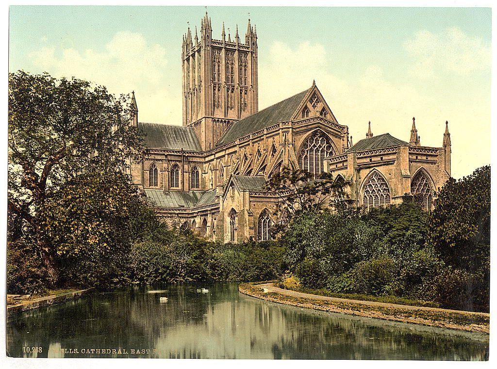 The image depicts a grand Gothic-style cathedral situated amidst lush greenery, with the majestic structure rising prominently in the background. The architectural features of the building include tall pointed arches, intricate tracery on windows and doors, buttresses supporting its massive walls, and large rose window adorning one section of the facade.

In front of the cathedral stands a tranquil pond reflecting part of the stately edifice and surrounding trees. A few ducks can be seen leisurely floating in the water, adding life to this serene scene. The foliage around the building is dense with various shades of green, indicating that it might have been taken during spring or summer.

The overall atmosphere exudes an air of historical significance and architectural beauty, capturing a moment where nature harmoniously coexists with human-made wonders. This image likely serves as an evocative representation of Gothic architecture in England, showcasing the aesthetic appeal and grandeur associated with such ecclesiastical buildings from that era.