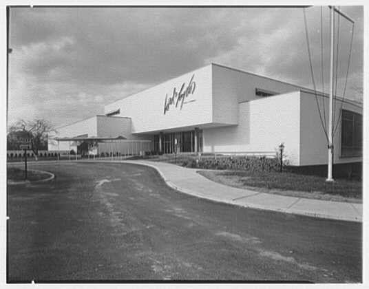 The image depicts the exterior of a large building with modern architecture, possibly from the mid-20th century. The structure has clean lines and appears to be two stories tall, featuring flat roofs without visible supports or ornamentation. A prominent sign on the facade reads "Lord & Taylor," indicating that it is likely associated with the department store chain known for its historical presence in retail.

The photograph is a black-and-white negative, suggesting an older vintage photo, possibly taken around 1955 as indicated by the caption text referencing "Exterior west facade" and "Gottscho-Schleisner." The company Gottscho-Schleisner was known for commercial photography in various industries.

The surrounding area of the building shows a paved road curving away from the entrance, with sidewalks on either side. There are street lamps lining one sidewalk, although no lighting is apparent as it might be daylight outside or simply not illuminated at that moment.

In summary, this image captures an exterior view of what seems to have been Lord & Taylor's store in Bala-Cynwyd, Pennsylvania during the 1950s. It provides a glimpse into early commercial architecture and urban planning for retail establishments from that era.