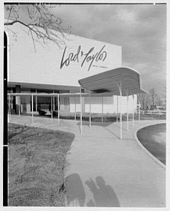 The image shows the exterior of a building with large, white letters that read "Lord & Taylor" prominently displayed on its facade. The photograph appears to be in black and white, suggesting it was taken some time ago. There is an overhanging roof structure extending from part of the building's front entrance. A paved walkway leads up to this entry area where two people can be seen walking away into a grassy field adjacent to the sidewalk.

The sky above seems partly cloudy with shadows visible on the ground, indicating sunlight filtering through clouds or possibly creating an artificial shadow effect due to lighting conditions in which the photo was taken. The architecture of the building has a modernist feel from its early design elements like large open spaces and clean lines characteristic of mid-20th-century commercial buildings.

The caption mentions "Lord & Taylor business," referencing this image being related to that company, while also providing information about Gottsch&Schneller Inc., which could be an entity involved in the creation or distribution of this photograph. The reference to a specific date suggests when the photo might have been captured - April 16th, 1955.

Finally, the source URL hints at where more detailed and possibly contextualized information about this image can be found online.