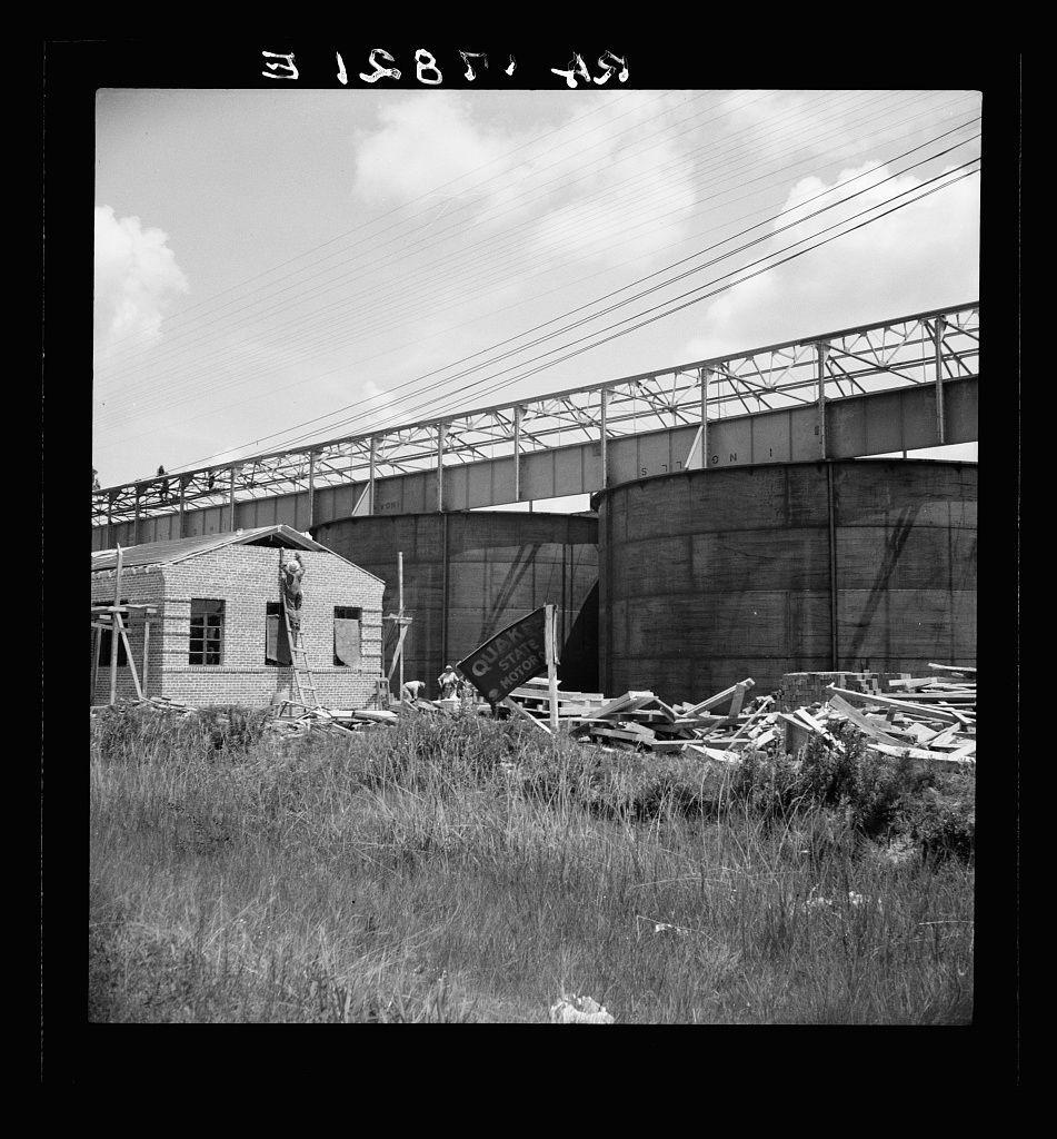 A black and white photograph depicting an industrial landscape. The focus is on a large, dilapidated turpentine still structure with visible rusting metal beams extending over the top of multiple cylindrical storage tanks labeled "STONER," suggesting they were used for storing processed turpentine. In front lies disarrayed lumber and debris near two smaller brick structures resembling old buildings or sheds; one appears to be in better condition, while another is collapsing into itself with its wooden framework exposed.

The foreground features tall grasses swaying gently under a sky dotted with clouds. The photo's monochromatic tone gives it an aged appearance, possibly from the mid-20th century, reflecting on industrialization and decline of small-scale processors in rural America during that era.