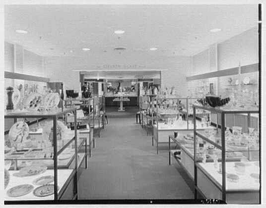 A black and white photograph of an interior setting, presumably a department store or shop selling various items. The layout consists of numerous shelves displaying dishes such as plates and bowls, arranged on the right side in what appears to be glass cabinets with doors. On the left side are also shelved displays containing similar dining ware. A person is partially visible at far end under display cases which possibly contain collectible or fine china.
The environment looks clean and well-lit by ceiling lights. The photograph does not clearly show any specific brand markings, but it captures a vintage aesthetic suggestive of mid-20th century retail design. Notable features include the abundance of dishware on sale, hinting at an emphasis on tableware in this particular store section or department.
Intriguingly, there is no direct indication that the photo relates to Lord & Taylor's business in Bala-Cynwyd, Pennsylvania; it appears unrelated based solely on visible elements. The date "1955 Apr 16" could be associated with the image but lacks contextual details within this description.
Further information provided includes a reference to Gottsch-Schleisner and an indication of one negative in size '4x5'.