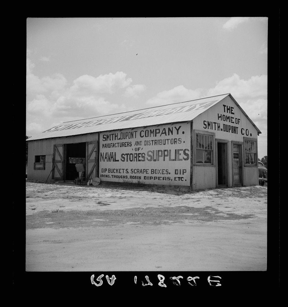 The image depicts a black and white photograph of an old, rural building labeled "THE HOME OF SMITH-DUPONT CO." It is identified as the headquarters for Smith Dupont Company. The structure appears to be part of their manufacturing operations or distribution center. Various products are listed on its side such as dip buckets, scrap boxes, dip irons, troughs, rosin drippers and other supplies.

This image seems like a historical snapshot capturing an industrial site in America during the early 20th century when small processors were prevalent for turpentine production. The text at the bottom of the photo suggests it is connected to large-scale industry practices involving agricultural products (turpentine) which was vital to American commerce and economy.

The photograph's age, its monochrome tone, along with some minor imperfections like scratches or stains on the film suggest that this image could be part of a larger archive documenting industrial history.