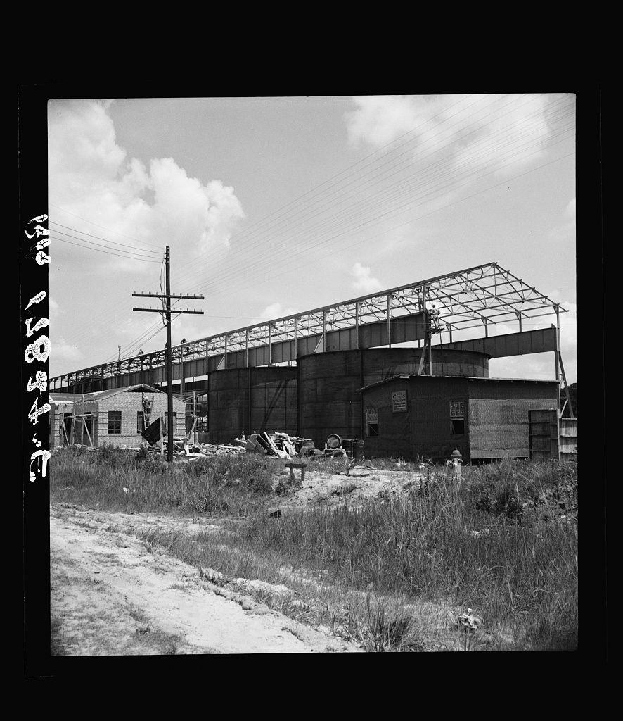 The image depicts a black and white photograph of an industrial scene, likely from the mid-20th century given its style. The main subject is a large structure with an exposed steel framework under construction or maintenance. It appears to be part of a larger complex that includes various smaller buildings, some in disrepair. In front of this central building are remnants of wooden structures and debris scattered across overgrown grassy terrain.

There's a sense of abandonment or temporary suspension of work due to the presence of these abandoned materials and unkempt surroundings. A utility pole stands prominently on the left side, with power lines extending towards it. The sky is partly cloudy but allows sufficient sunlight that illuminates the scene unevenly. There are no visible workers or machinery in operation, adding a stillness to the image.

The photograph seems to capture an industrial setting possibly related to processing plants as indicated by historical references mentioning turpentine and small processors from Georgia. This could suggest it's part of a series documenting industry-related landscapes during that era.
