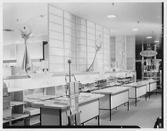 The image depicts an interior view of a fashion linen counter at Lord & Taylor, located in Bala-Cynwyd, Pennsylvania. The setting appears to be from the year 1955 on April 16th and is captured as part of a collection by Gottscho-Schleisner, Inc., which specializes in photographic negatives.
In this photograph, we observe several long counters stretching across the frame, with various pieces of linen or fabric displayed for sale. The space seems to be organized neatly, featuring shelves stocked with folded items and tables topped with flat linens. Decorative elements such as flower arrangements can be seen placed strategically throughout the area. Above, fluorescent lighting fixtures illuminate the merchandise.
The overall atmosphere is clean, orderly, and indicative of mid-20th-century retail design aesthetics. The monochromatic nature of this image suggests that it was taken during a time when color photography was not yet widely accessible or commonly used in commercial settings.
