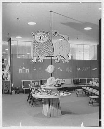 The image shows a black and white photograph of an interior space, likely a retail store or public building. In the center is a large decorative piece hanging from the ceiling, featuring cartoon-like figures such as owls, stars, planets, and other celestial objects in a whimsical style. Below this decoration are several round tables with chairs arranged around them, suggesting a communal area for children to play or gather.
The background reveals more of the interior design, including what appears to be shelving units against the wall on one side and benches lined up parallel to it on another side. The overall atmosphere is playful yet orderly, evoking an impression from mid-20th century retail establishments that catered specifically towards children's needs or interests.
The image seems to come with additional context indicating its source: a business in Bala-Cynwyd, Pennsylvania called Lord & Taylor; it mentions "Lion in Children's Shoes" and the date April 16, 1955. This contextual information suggests that this photograph is part of an archive documenting various items or scenes related to retailing during that era.
The image carries a unique identifier (Gottscho-Schleisner) associated with Gottscho-Schleisner Inc., which might be the photographer, photo studio, or organization responsible for preserving and cataloguing these images. The format of one negative in saf [...]