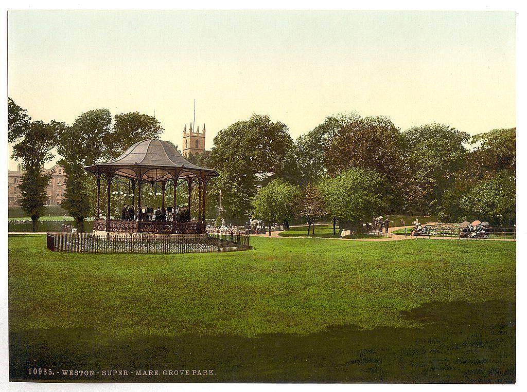 The image shows a vintage postcard depicting an outdoor scene in the park. In the foreground, there is a well-maintained grassy area with a prominent bandstand structure featuring ornate metalwork and a pointed roof supported by decorative columns. The bandstand appears to be constructed of wood and has benches around it for visitors.

In the middle ground, several people can be seen enjoying the park. Some are sitting under parasols on tables arranged in rows along pathways that wind through the greenery. Others stroll leisurely or rest against trees.

The background reveals a mix of mature trees providing shade to various sections of the park and glimpses of historic architecture, including what looks like an ornate church spire with Gothic design elements atop its tower.

Overall, this image captures a serene moment in Weston-super-Mare's Grove Park during early 20th-century England.
