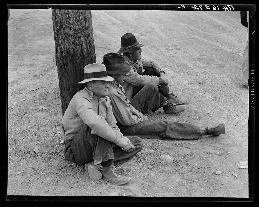 The image depicts three men seated on the ground beside a tree trunk. They appear to be in an outdoor, possibly rural setting with bare earth and small rocks visible around them. All three are wearing hats; two of whom have their feet propped up against what seems like a utility pole or large log. Their clothing is modest and worn-looking, suggesting practical work attire rather than leisurewear. The men's expressions are serious, and they seem to be in contemplation or waiting for something.

The photograph has the distinct graininess characteristic of early 20th-century black-and-white film photography. There is handwritten text on the photo border which reads "Semi Monthly Relief Checks," indicating that these individuals may be among those receiving financial assistance from a relief program, possibly related to economic hardships such as unemployment or drought during the Great Depression era.

The image captures a moment of quiet resilience and waiting in difficult times, reflecting broader social issues at play during that historical period.