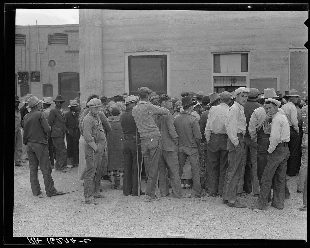 The image depicts a large group of people gathered outside what appears to be an administrative building, likely during the Great Depression era. Individuals are standing in line or huddled together, wearing varied attire such as suits, hats, and overalls indicative of working-class clothing from that period. Many wear hard hats typical for manual laborers at construction sites. The scene is outdoors with a clear sky above; it's daytime but no shadows suggest the sun isn't directly overhead, possibly indicating morning or late afternoon light conditions.

The building in the background has a plain facade and features such as windows without glass panes, hinting at an institutional setting for dispensing aid like relief checks. The ground is unpaved with visible dirt patches, reinforcing the economic hardship context of the time. There's little vegetation around, further emphasizing arid environmental conditions.

The photograph itself carries historical significance; it seems to capture a moment where people are waiting in anticipation or anxiety, likely anticipating receiving their semi-monthly relief checks as mentioned in the caption from Calipatria, California. The black and white nature of the image underscores its age and contributes to the somber mood conveyed by both content and composition.

The photograph is credited with text that includes a location "Calipatri [...]