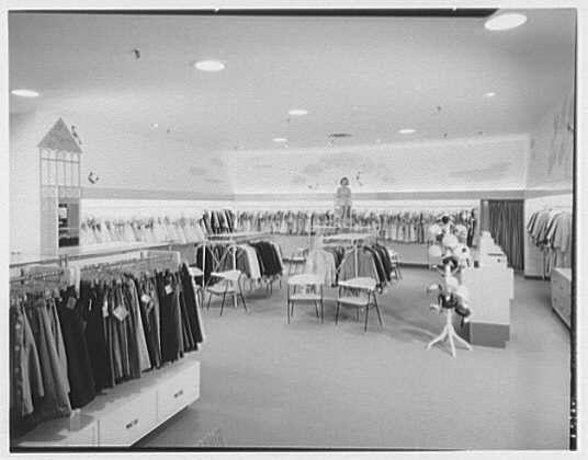 This black and white photograph depicts a spacious girls' clothing department inside the Lord & Taylor business in Bala-Cynwyd, Pennsylvania. The image shows rows of garments hanging on racks along both sides of an open floor area, which is illuminated by overhead lighting fixtures. A variety of women's apparel including coats, dresses, and jackets can be seen displayed prominently for customers to browse through.
In the center of the room stands a woman wearing glasses, who appears to be shopping or possibly browsing merchandise. There are additional clothing racks with more garments in the background towards the far end of the store. The atmosphere suggests an organized and well-lit retail environment typical of mid-20th-century American fashion stores.