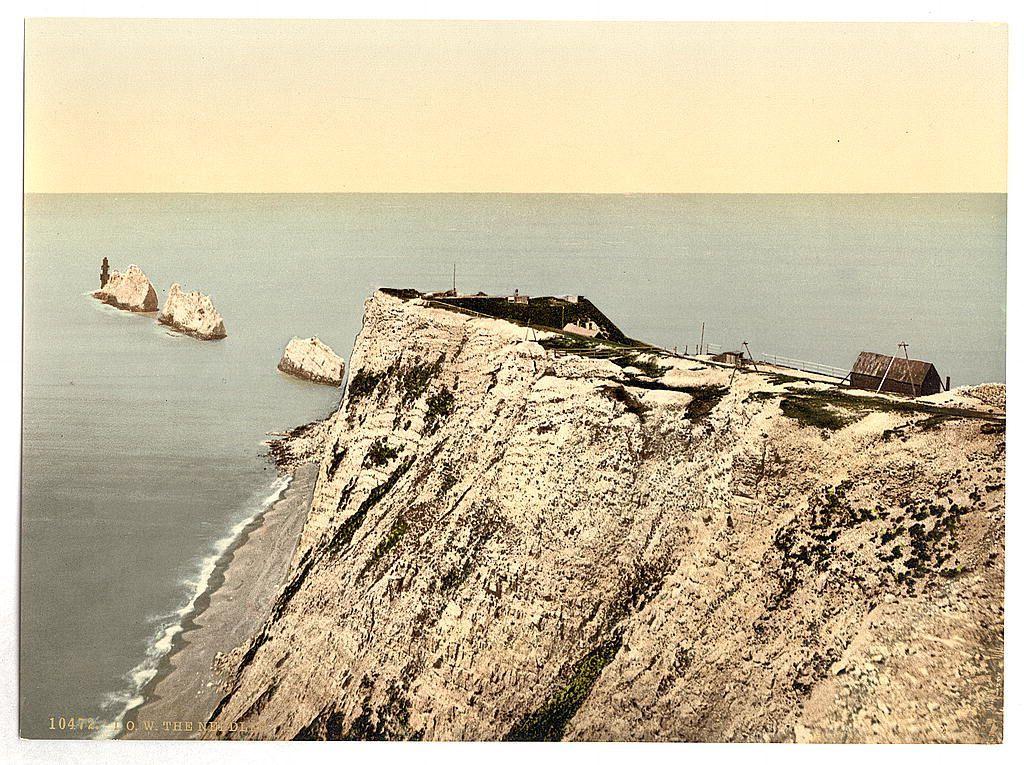 This image depicts a coastal scene featuring the famous chalk formations known as The Needles on the Isle of Wight in England. These natural rock pillars extend vertically out of the sea, creating an impressive geological feature visible from various points along the coastline.

In this particular photograph, we can observe one prominent cliff with structures that appear to be part of a historical defense installation or lighthouse facilities. A few individuals are seen on top and around these structures, giving scale to their size against the vastness of the surrounding environment.

The coloration suggests an early print from the late 19th century, capturing the natural beauty and human presence in harmony with this coastal landscape. The calm waters reflecting a hazy sky add tranquility to the scene while highlighting the dramatic cliffs' rugged texture.

This image serves as valuable historical documentation of Britain's landscapes during that era and offers insights into maritime activities associated with such landmarks.