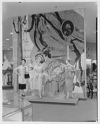 The image is a black and white photograph depicting an interior scene, likely from a vintage department store. The setting features three female mannequins dressed in mid-20th-century fashion attire, standing on what appears to be a raised platform or podium within the storefront display window.

These mannequins are posed with their arms outstretched and hands positioned near each other as if engaging in conversation, creating an interactive visual dynamic. The clothing displayed includes various styles of dresses from that era, such as knee-length skirts, blouses, cardigans, and accessories like hats or handbags.

In the background within the display window is a mural with intricate details featuring what seems to be stylized botanical elements resembling roots or vines intertwined in complex patterns. Also visible are two large cutouts depicting female figures wearing suits; these may represent coats and jackets available for sale at the store, possibly during an April promotion as indicated by the caption.

The overall composition of the image suggests a promotional display designed to attract customers' attention with its thematic elements, clothing styles, and visual merchandising techniques characteristic of that period. The photograph's historical context is further supported by information provided in the reference source mentioned at the end, which indicates it  [...]