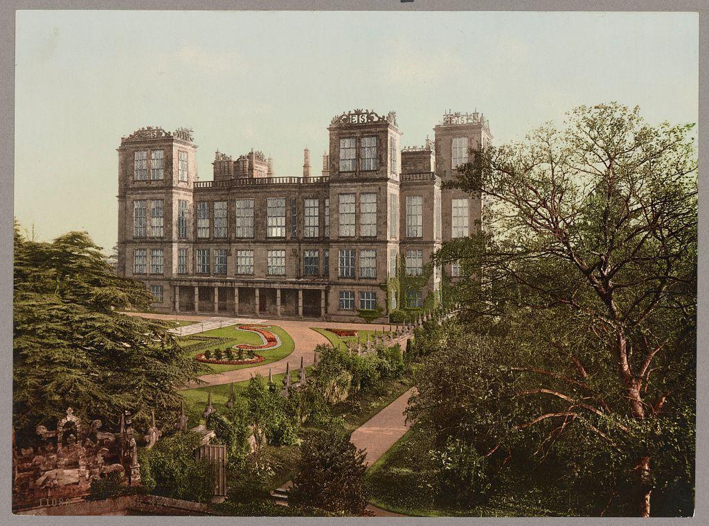 This image depicts a grand, multi-level historical building with intricate architectural details. The structure features multiple windows and ornate decorations on its facade, including sculptures or reliefs at the corners of each level. A well-manicured garden surrounds the estate, with curving pathways leading towards it from different angles within what appears to be an expansive parkland setting. Trees are scattered throughout the landscape, providing a lush green canopy over parts of the grounds and contrasting against a clear sky in the background. The overall atmosphere conveyed by this image suggests opulence and grandeur associated with historical estates or palatial residences.