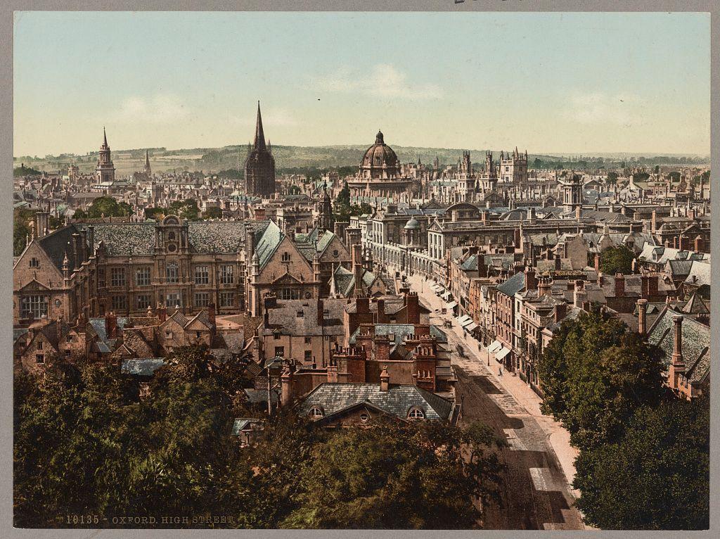 The image portrays a bird's eye view of Oxford, England during the 19th or early 20th century. It is an aerial perspective showcasing High Street and surrounding areas in great detail.

The architecture dominates with numerous historic buildings featuring steeply pitched roofs, ornate facades, chimneys, and varied window styles indicative of Victorian design. The street layout suggests a dense urban fabric typical for the period. In the distance are prominent landmarks such as spires, domes, towers, and other notable structures suggesting religious or institutional significance.

Trees line parts of High Street, adding greenery to an otherwise bustling town scene. There's no visible movement on the streets which could imply this photo was taken at a quiet time during daily life in Oxford. The coloration leans towards sepia tones common for photos from that era, giving it a historical feel.