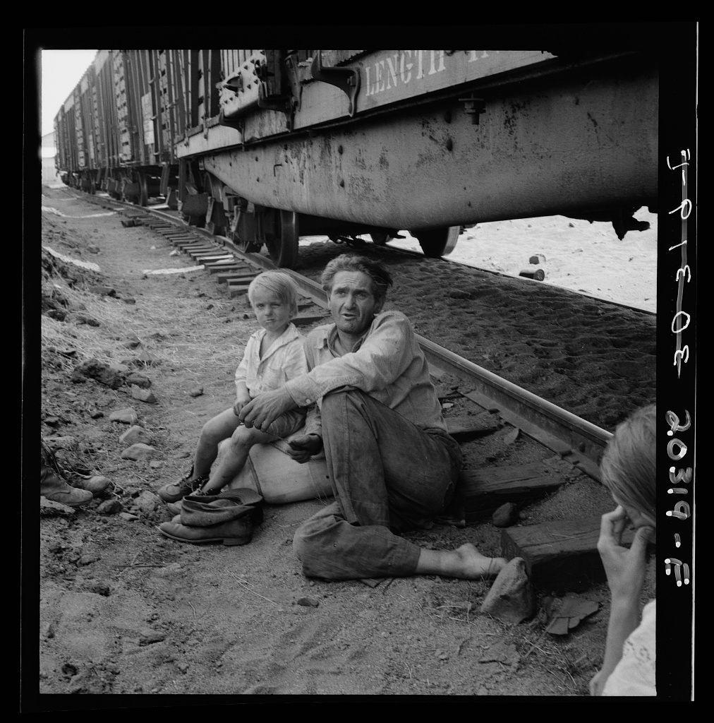 The image depicts a poignant moment in black and white. Two individuals, possibly father and child based on their close proximity and body language, are seated on the ground beside railway tracks. The adult is reclining with legs crossed at the ankles, wearing a shirt that appears worn from use or hardship. His right leg extends out while his left rests comfortably against an armrest made of wood slats.

Beside him sits a child, possibly in their early years given their size and stature compared to the seated figure. The child is barefooted, with one shoe resting on top of what seems like a sock or rag beside them. Their legs are crossed as well, mimicking an adult posture yet maintaining innocence through the simplicity.

In front of these two figures lies another individual's hand in close proximity, possibly indicating someone else being part of this group but not fully visible within the frame. The sand beneath their feet suggests they may be near a beach or similar environment with loose soil.

The backdrop reveals railway tracks leading into the distance where freight cars are stationed parallel to them, suggesting an abandoned or less-frequented section of the track system. There is also debris scattered around indicating neglect and disrepair in this area. The overall setting speaks volumes about their circumstances hinting at displacement, hardship, possibly migr [...]