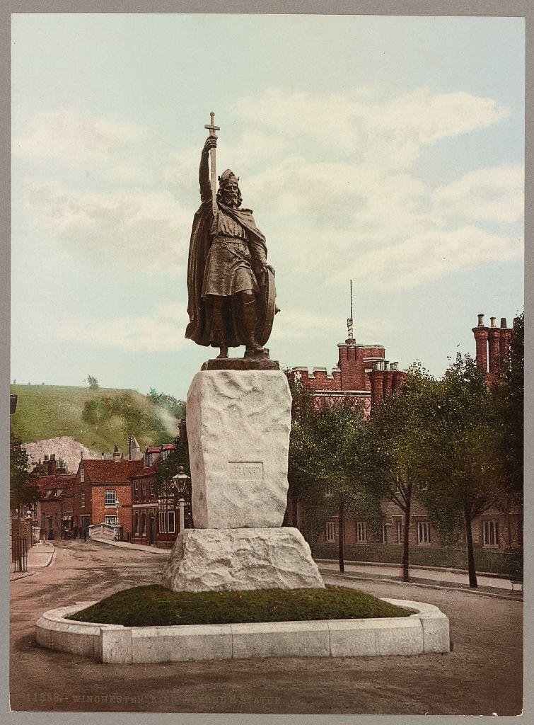 The image shows a bronze statue of King Alfred the Great standing on an elevated stone pedestal. The king is depicted holding a sword in his right hand, with one arm raised as if delivering a speech or rallying troops. He wears traditional English attire from that era: a long robe and a cloak with fur trimmings around the edges. His headgear appears to be of royal status, possibly indicating his noble lineage.

The statue is placed within an urban setting; in the background, there are buildings reminiscent of 19th-century architecture, including red brick structures with multiple windows. A street runs along one side of the pedestal, and a small green space surrounds its base. The sky above appears partly cloudy, suggesting it might be either morning or late afternoon.

The overall tone of the image suggests historical significance, capturing an iconic representation of King Alfred in his adopted city's public space. This statue commemorates the king who was instrumental in establishing English law after Viking invasions and is considered a key figure in British history for laying down some laws on which England was based as well as preserving much of Anglo-Saxon culture.

The image has visible text, "1884- WINCHESTER KING ALFRED ON EARTH'S STAND," indicating the date it was possibly taken or published and its subject matter.