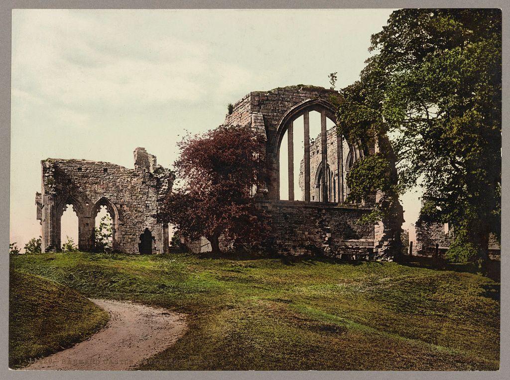 The image depicts the ruins of Eggleston Abbey located in Durham, England. The historical structure features Gothic architectural elements such as large arched windows and stone walls that show significant decay over time. In the foreground, there is a grassy area with patches of sunlight filtering through trees, creating dappled shadows on the ground. A dirt path meanders from the bottom left corner into the ruins, inviting exploration. The sky appears partly cloudy, suggesting an outdoor setting during daylight hours. This photochrom print likely dates back to around 1890-1906 and captures the serene yet haunting beauty of these historical remains amidst nature's reclamation.