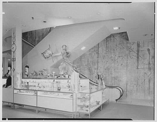 The image depicts an indoor scene, likely within a department store or exhibition space. A prominent display case filled with various items is situated in the center foreground. It features glassware and decorative objects arranged on shelves against white backdrops.

Behind this main exhibit, there's a larger structure that appears to be part of a themed installation or architectural design element. This feature consists of an arched opening leading into another space where figures are posed within what seems like classical attire, suggesting historical costumes or characters from different eras being showcased in the display case on the left.

The overall setting includes multiple shelves with items displayed at various heights and angles, creating a dynamic presentation for viewers navigating through this area. The architecture above has smooth curves, indicating modern design principles of that era. Overall, it conveys an atmosphere typical of mid-20th-century retail environments or exhibition spaces dedicated to cultural artifacts.

The image bears the timestamp "Fantasia II 1956 Mar. 6," suggesting its connection with a specific event held on March 6, 1956, possibly linked to Lord & Taylor business in Garden City, Long Island as mentioned elsewhere. Gottsch-Schleisner is credited for creating this photograph, capturing the moment during an exhibition or promotional  [...]