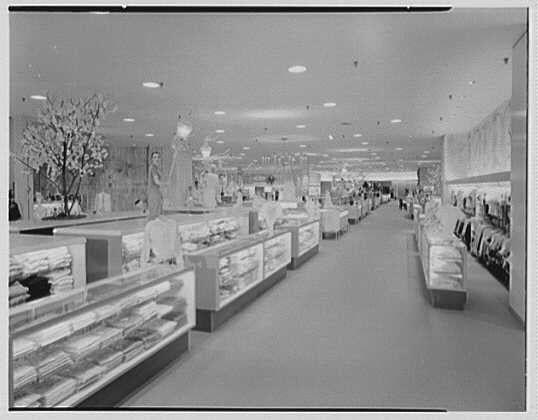 The image is a black and white photograph depicting the interior of what appears to be an upscale department store, likely from the mid-20th century. The perspective shows a long aisle leading through the center of the establishment, flanked by display cases containing various items such as jewelry or watches on the left side and clothing racks with mannequins dressed in fashion attire on the right.

The ceiling is adorned with multiple round lights that provide illumination to the space. On either end of the central aisle are larger display areas showcasing more products; one displays a large, lit floral arrangement at its entrance while another has models posed as if shopping or browsing items within it. 

Various signage and advertisements can be seen throughout, contributing to an atmosphere typical of mid-century retail environments aimed at creating an inviting ambiance for shoppers.

The overall impression is that of a well-maintained, bustling department store designed with attention to aesthetics and customer experience during its era.