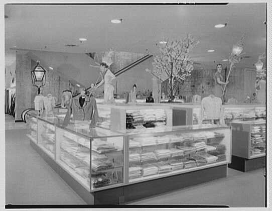 The image depicts an interior scene of a retail store, likely from the 1950s era. The photograph is in black and white and shows various clothing items displayed on shelves within glass cases. There are at least two people visible: one appears to be a shopper or salesperson holding a broom near some plants with blossoming branches, while another individual seems to be interacting with the merchandise. A staircase leading up possibly indicates multiple levels of store floors.

The environment suggests a well-lit and organized retail space typical for that time period. It's possible this photo was taken at Lord & Taylor business in Garden City on March 6, 1956, as mentioned in additional information provided about the image source from Gottschl-Schleisner Inc., an entity associated with archival photographs.

The photograph has a vintage quality and captures both functional elements of retail display such as clothing racks and shelving units alongside decorative items like plants. The overall setting gives insight into shopping aesthetics, store layouts, and merchandise presentation during that era in the United States.