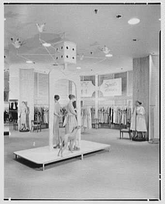 The image depicts a vintage retail store, likely from the mid-20th century. Several individuals are seen inside the establishment; three people stand on raised platforms in what appears to be an advertisement or display setup for clothes and accessories. The attire of these figures suggests they might represent mannequins dressed in fashion garments.
The background features multiple racks filled with clothing, presumably hanging items meant for sale. There's a sense of spaciousness within the store as indicated by its high ceiling adorned with various lights fixtures which cast illumination throughout the space. Overall, this photograph captures an era where shopping experiences were more theatrical and attention-grabbing compared to contemporary settings.
In terms of additional context, it is mentioned that Lord & Taylor was located in Garden City on Long Island during March 6th, 1956. Gottschalk-Schleisner Inc., potentially a photography or archival company associated with this image, likely played a role either as the photographer or custodian responsible for preserving such historical images. The reference to 'Dovecote' could indicate another related term or project connected to these entities.