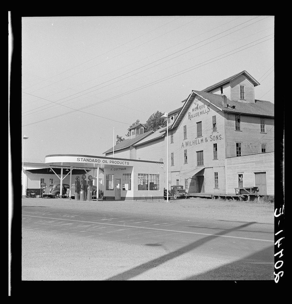 The image is a black and white photograph depicting an old-fashioned gas station next to the Monroe Roller Mills, which appears as "A Wilhelm & Sons" mill. The scene looks like it could be from mid-20th century America given its style of architecture, vehicles visible at the pump area, and overall aesthetic. There's no explicit information about this particular location being in Oregon or Benton County within a provided caption that I can discern without additional context. This photograph appears to show an industrial site with historical significance due to the presence of milling mills which were often key parts of rural economies during earlier decades before roads improved transport, making them hubs for local commerce and community gatherings as implied by the accompanying text on the image.