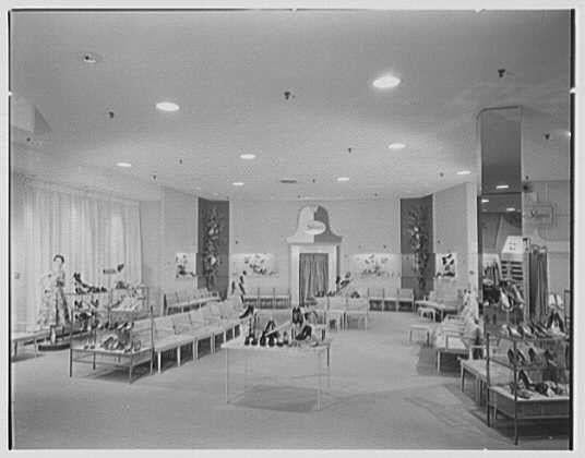 The image shows an interior view of a shoe store, likely from the 1950s era based on its vintage appearance. The layout is spacious and well-lit with several display cases filled with various styles of shoes arranged throughout the room. There are at least four large display windows showcasing mannequins dressed in different attire to complement the footwear being sold inside.

The store features a mix of seating options, including benches for customers seated on one side, tables presumably for trying or examining shoes closer up, and chairs placed near other sections. The design includes decorative elements like wall-mounted displays with more products, possibly featuring posters or advertisements relevant to that time period's fashion trends.

Overall, the atmosphere appears orderly and clean, catering towards a comfortable shopping experience.