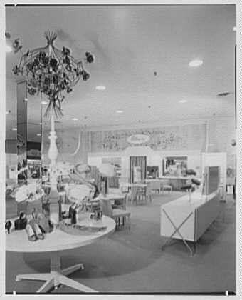 The image depicts an interior space, likely a department store or showroom. The scene is dominated by furniture and decorative items on display. A large table with a white surface occupies the center of the room, surrounded by chairs and other seating arrangements. Several lamps are placed around the room, providing illumination to highlight various objects.

On one side of the room, there's an eye-catching chandelier hanging from the ceiling, featuring numerous bulbs arranged in a decorative pattern. The walls appear adorned with artwork or mirrors, adding visual interest to the space.

In terms of color scheme, it is difficult to determine as the image appears black and white, which suggests that this photo was taken during an era when full-color photography wasn't common yet. The overall atmosphere conveyed by the photograph has a vintage feel, likely from the mid-20th century based on the furniture styles.

The caption also includes information about Lord & Taylor's business in Garden City, Long Island and mentions Gottschalk-Schleisner Inc., which could be related to photography or marketing. The reference number '691f' might indicate an archival cataloging system for photographs within a collection at a particular institution or individual collector.

In summary, the image captures a well-appointed display area with furniture and lighting fixtures on show in what se [...]
