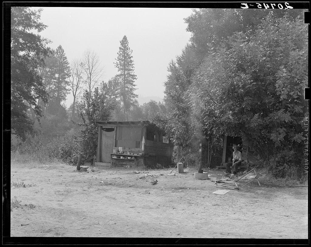 The image displays an old black and white photograph depicting a rural setting. In the center, there is a makeshift wooden structure with visible windows and doors that appears to be serving as living quarters or storage space. Around it are scattered various objects such as pots, mugs on stands, pieces of wood, and what seems like debris on the ground indicating an unkempt environment.

To the right side of this central shelter, there is a smaller wooden structure with its front facing outward; someone appears to be seated inside at a makeshift table. This person could possibly be engaged in some activity such as eating or working over something placed on the tabletop.

Surrounding these structures are trees and shrubs that give an impression of being situated within a forested area, indicating perhaps this setting is located off-the-beaten-path or not well-maintained. The overall atmosphere suggests hardship, temporary living conditions, possibly during early to mid-20th century judging by the style of shelters.

The photograph's clarity and monochrome tones give it an aged appearance with soft focus on distant objects, hinting at either a long shutter speed used while capturing this photo or possible degradation over time.