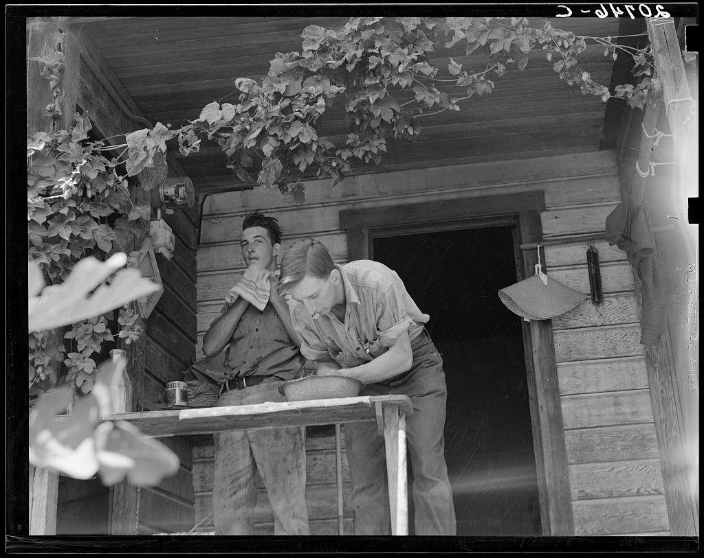 The image depicts a black-and-white photograph featuring two individuals on a porch. The scene appears to be set in an old house with wooden siding and lattice work, indicative of rural or farm-like surroundings. One individual is standing behind the other who is bent over at a makeshift table, seemingly engaged in some task that involves handling objects placed atop it.

The setting features lush foliage cascading from above, hinting at a garden or vineyard nearby. Various items such as bottles and possibly agricultural tools are visible on the porch surface, suggesting domestic life intertwined with work-related activities. The attire of both individuals suggests practical, utilitarian clothing typical for farm labor during an earlier era.

The photograph's grainy texture and monochrome palette contribute to its historical atmosphere, potentially dating it from a period in which such imagery was prevalent. Given the visual cues - rural setting, domestic scene, old-fashioned dress code – this image likely captures daily life within a farming family or community associated with agriculture during an earlier time.

This photograph is part of Oregon's rich agricultural history and represents a slice-of-life that offers insight into the lived experiences of individuals connected to hop farming. The image carries historical significance as it encapsulates elements from rural  [...]