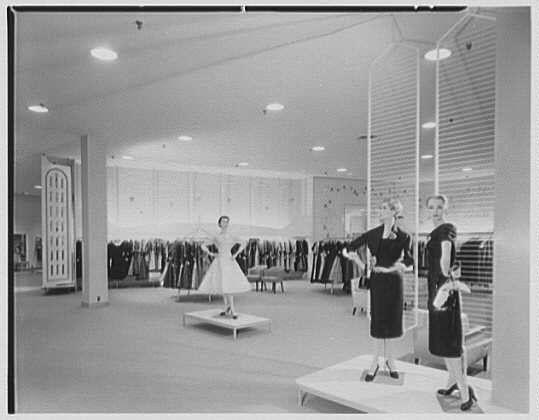 The image depicts a vintage clothing store with three women standing on platforms displaying fashion garments. The primary woman, in the center-right of the frame, is dressed in a dark knee-length dress and holds a garment over her shoulder. To her left stands another female figure in a black outfit also holding an item to display. On the far right, there's yet another person wearing similar attire.

In the background, rows upon rows of dresses hang on racks against wall-mounted fixtures lined up with horizontal slats for illumination. The space is brightly lit by ceiling lights and appears organized and spacious as it extends towards a large windowed area or possibly an entrance to the building in which this store operates.

The image has a grainy texture typical of black-and-white photographs from mid-20th-century America, enhancing its historical context. There's no explicit indication regarding race or gender due to the fashion norms and photographic technology at that time period; it portrays women working within their professional setting during what appears to be an advertisement for Lord & Taylor in Garden City on March 6, 1956.