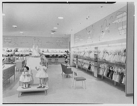 The image depicts an interior view of a department store, specifically focusing on the children's and boys' clothing section. It is in black and white, suggesting it was taken during a time when color photography was less common or not used for promotional materials.

In the foreground, there are two mannequins dressed in early 20th-century style dresses: one appears to be wearing pinafore-type garments while standing on a small platform. Behind them is another display featuring an array of children's and boys' clothing hanging neatly on racks against a wall adorned with decorative designs or illustrations.

The store has bright lighting, likely from overhead fluorescent lights, creating reflections visible on the shiny floor surface in front of the mannequins. In addition to the main walkway seen here, there is another aisle that seems to lead deeper into the store where more clothing options might be available for exploration by customers or shoppers.

The composition and layout suggest a well-organized retail environment designed with aesthetics as much importance as convenience for shopping purposes. The absence of modern digital devices indicates this photo was taken before such technology became ubiquitous in stores, emphasizing personal customer service interactions likely to have been prominent during that era.