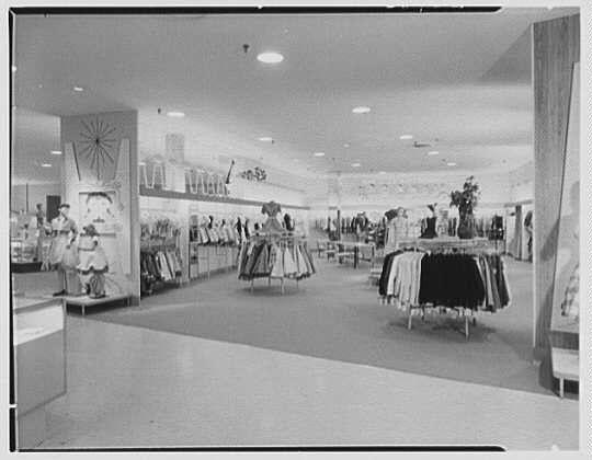 The image depicts an interior view of a department store, likely from the mid-20th century based on fashion styles and layout. Rows upon rows of clothing are displayed in various sections: dresses suspended by stands to one side, skirts hung together, and what appears to be outerwear or coats neatly arranged at another section.

The flooring is polished wood with visible grout lines between tiles, suggesting a well-maintained commercial space designed for easy cleaning and movement. The lighting comes from overhead fixtures that are typical of stores in mid-20th-century America - large circular lights embedded into the ceiling panels to illuminate merchandise below.

In terms of organization within the store, it is clear that there's an emphasis on categorizing clothing items by type; dresses, skirts, coats or jackets all have their designated areas. This kind of layout was common in department stores as it allowed for easy customer navigation and quick identification of desired products based on category.

The overall ambiance seems to be quite spacious with ample room between displays which could suggest a larger store size than what's typical today due perhaps to the design standards or consumer expectations from that era regarding shopping space.