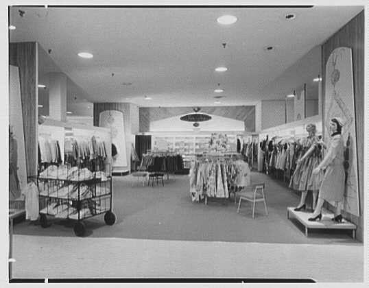 The image depicts an interior view of a vintage clothing store, possibly from the 1950s. The setting is well-lit with overhead lights illuminating the space. On one side, there's a display rack filled with various articles of clothing hanging on hangers and shelves below it containing folded garments or accessories.

The center area features rows of dresses hung on racks for customers to browse through, showcasing an array of styles and lengths. To the right stands a mannequin dressed in what appears to be a light-colored dress with details that suggest mid-century fashion trends. The store has large floor-to-ceiling mirrors reflecting part of its interior.

The overall atmosphere is reminiscent of a bygone era's shopping experience, characterized by organized displays, ample lighting, and an inviting ambiance for shoppers interested in women’s apparel from the specified time period.