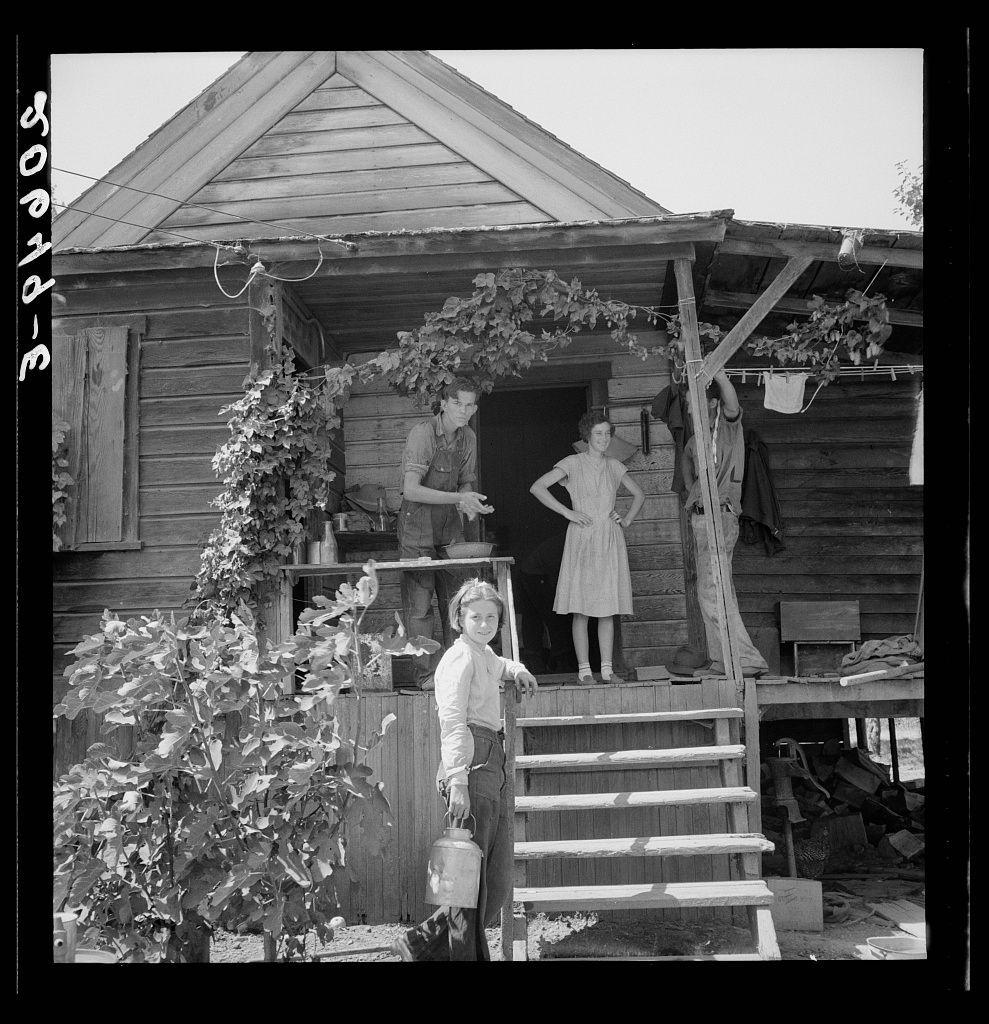 The image is a black and white photograph depicting a rural scene. It features the front porch of a wooden house with steps leading up to it, adorned by climbing plants on one side. On this porch, two children are present; a boy in overalls carrying what appears to be milk cans or similar containers, and another child dressed in light clothing standing beside him.

In the background stands an adult man wearing work clothes as he is seen working with some machinery connected to a pole extending upwards from the house's roof. The environment suggests agricultural activity, likely related to hop farming given the context provided by text overlay that reads "Hop farmer’s children, small owner, backyard of house." This indicates that these individuals are part of a family involved in hop farming.

The setting is identified as Oregon, specifically Polk County near Independence based on additional information. The photograph captures an authentic moment from rural life during what could be the mid-20th century given its style and content.