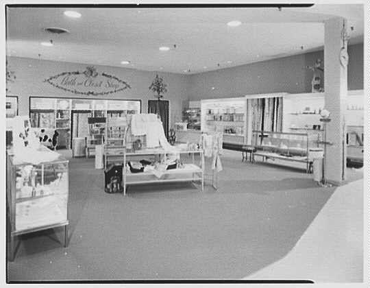 The image depicts an interior view of a retail store from the mid-20th century. The setting appears to be a "Bath and Closet Shop" as indicated by the signage on the wall at the far end of the room, suggesting that it specializes in clothing or personal hygiene products.

There is a large display with various items draped over what seems like an ironing board or a garment rack. Several shelves filled with folded clothes are visible around this central area. On one side, there's a counter with glass displays showcasing boxed goods, likely related to the store’s offerings.

The flooring appears carpeted, and overhead fluorescent lighting illuminates the space. The room is largely empty of people, providing an unobstructed view of the merchandise arrangement. In the background on the right-hand side, there are more shelves or display cases with items neatly arranged, extending towards a wall that features additional signage.

The image's monochromatic color scheme and style give it a historical feel, characteristic of black-and-white photography from around 1956 as mentioned in the context provided. The absence of modern elements like digital screens or contemporary marketing materials suggests this photo captures an earlier era of retail environments.