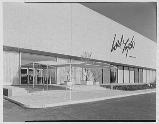 The image depicts a black and white photograph of the exterior facade of Lord & Taylor, located in Garden City. The building has an expansive design with wide-open entrances that have canopied areas over them for shelter. There are several large glass doors leading into what seems to be multiple levels or departments within the store.

In front of the entrance stands a set of concrete steps flanked by two short walls and potted plants, adding some greenery to the otherwise stark setting. The structure itself is characterized by clean lines with visible horizontal bands that break up its flat expanse. A prominent feature on this side facade is the "Lord & Taylor" sign, written in cursive script above a row of rectangular windows.

The photograph appears vintage and likely dates back to March 9th, as noted at the bottom right corner indicating it was taken by Gottschol-Schleisner Inc., suggesting that it might be from an early period for Lord & Taylor's business operations or architecture.