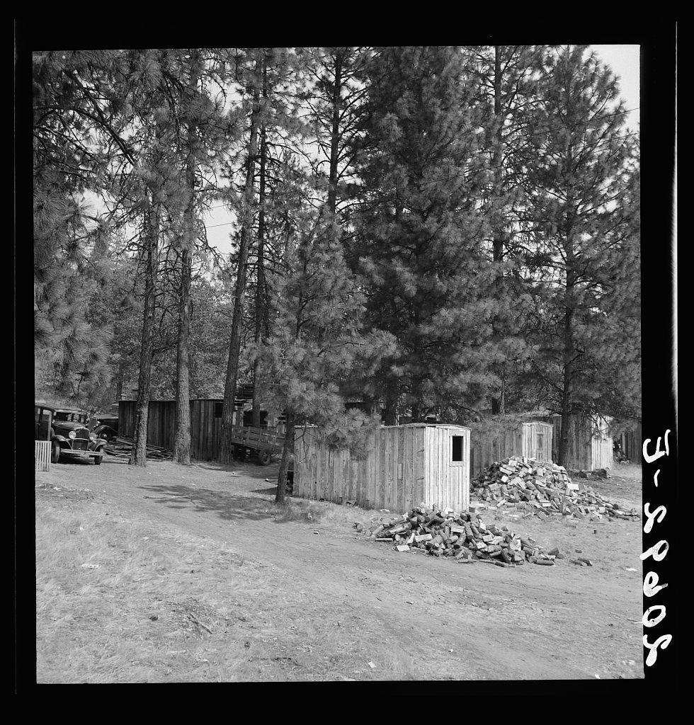 The image is a black and white photograph depicting an outdoor scene with tall pine trees dominating the background. In front of these trees, there are several small wooden cabins or sheds situated on what appears to be undeveloped land with sparse grass coverage. A pile of discarded bricks or stones lies near one of the cabins, indicating possible construction work or abandonment. Behind the structures and along a dirt path, old cars can be seen parked, suggesting this might be an area for storage or perhaps once used as living quarters in a rural setting.

The overall atmosphere is rustic, with a sense of quietness typical of remote areas away from urban settings. The absence of water sources nearby suggests that it could be located within arid conditions where rainfall and runoff are sparse enough to not support extensive vegetation growth around the structures.