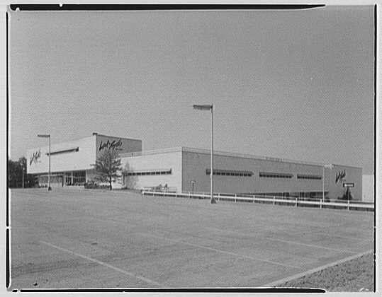 The image depicts a large, modern shopping center with multiple buildings and parking lots. The main structure has the Lord & Taylor name prominently displayed on its facade. A few smaller outbuildings are connected to this central building by what appears to be covered walkways or corridors. There is an open lot in front of the complex, suggesting it serves as a primary entrance area.

The photograph is presented in black and white, which provides contrast against the buildings' muted colors. The sky above seems clear with no visible clouds. Street lamps are positioned sporadically around the parking lot but none are illuminated or active at this time captured by the camera's lens. There isn't any discernible movement within the scene; it exudes a stillness, perhaps indicative of an off-peak hour.

The caption indicates that the photo relates to Lord & Taylor in Bala-Cynwyd, Pennsylvania, and notes its entrance on Belmont Ave., dated June 7th, 1956. The image is credited to Gottschlo-Schleisner Inc., suggesting it may be a professional architectural or construction photograph.

Overall, the photo presents an early depiction of what could have been one of America's more modern shopping centers during its time captured in this snapshot from June 7th, 1956.