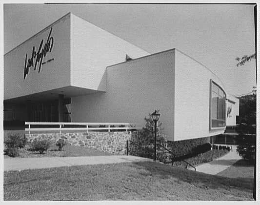 This black and white photograph depicts the south facade of a building from 1956, which is identified as Lord & Taylor. The structure features clean lines with minimalistic architecture that includes large windows on one side. There's signage prominently displayed across the top left corner of the building bearing the company name in stylized lettering. In front of this modern-looking commercial facility stands a lamppost and it appears to be set within landscaped grounds, as grass is visible alongside some rocks or stones at its base. This image has an accompanying caption that reads "Lord & Taylor business Bala-Cynwyd Pennsylvania South facade sharp 1956 June 7" with additional information indicating the photographer was Gottscho-Schleisner, Inc., and mentioning a negative size of safety;4x5in. The source for this image is https://images.loener.nl/gottscho-schleisner/full/691f/691f0231450e57a267505169.jpg