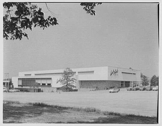 The image depicts a black and white photograph of the Lord & Taylor business located in Bala-Cynwyd, Pennsylvania. It is an afternoon view from June 7, 1956, as mentioned by Gottscho-Schleisner, Inc., who took this photo.
In the foreground, there are some leafless tree branches partially framing the top left corner of the image. The main focus lies in a large building with "Lord & Taylor" prominently displayed on its facade. This one-story structure has numerous windows and appears to be a spacious commercial establishment during that era. In front of the store, several cars are parked or driving by, indicating some activity around this business.
The ground is mostly covered with asphalt, suggesting ample parking space for patrons visiting Lord & Taylor. The sky seems clear without any visible clouds, casting even lighting across the scene and accentuating the details on the building's facade. This image provides a glimpse into retail architecture and urban development from the mid-20th century in America.
The source of this photograph is Gottscho-Schleisner, Inc., with reference to one negative that measures 4x5 inches (13 cm x 13 cm). The image can be found on their website at: https://images.loener.nl/gottscho-schleisner/full/691f/691f0232450e57a26750516f.jpg.