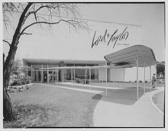 The image depicts a black and white photograph of the exterior facade of Lord & Taylor's department store located in Bala-Cynwyd, Pennsylvania. It appears to be from June 7th, 1956, as indicated by the caption mentioning "West entrance I" with that date. The building is modernist architecture characterized by clean lines and minimalistic design elements like large glass panels on the facade, supported by white metal frames. A prominent awning extends over a walkway leading to the main entrance where a solitary figure can be seen walking past. Trees are visible in front of the store's lawn area, with one particularly noticeable tree trunk extending into the frame from the left side. The iconic "Lord & Taylor" signage is prominently displayed above the building entry, stylized elegantly against the clear sky backdrop.