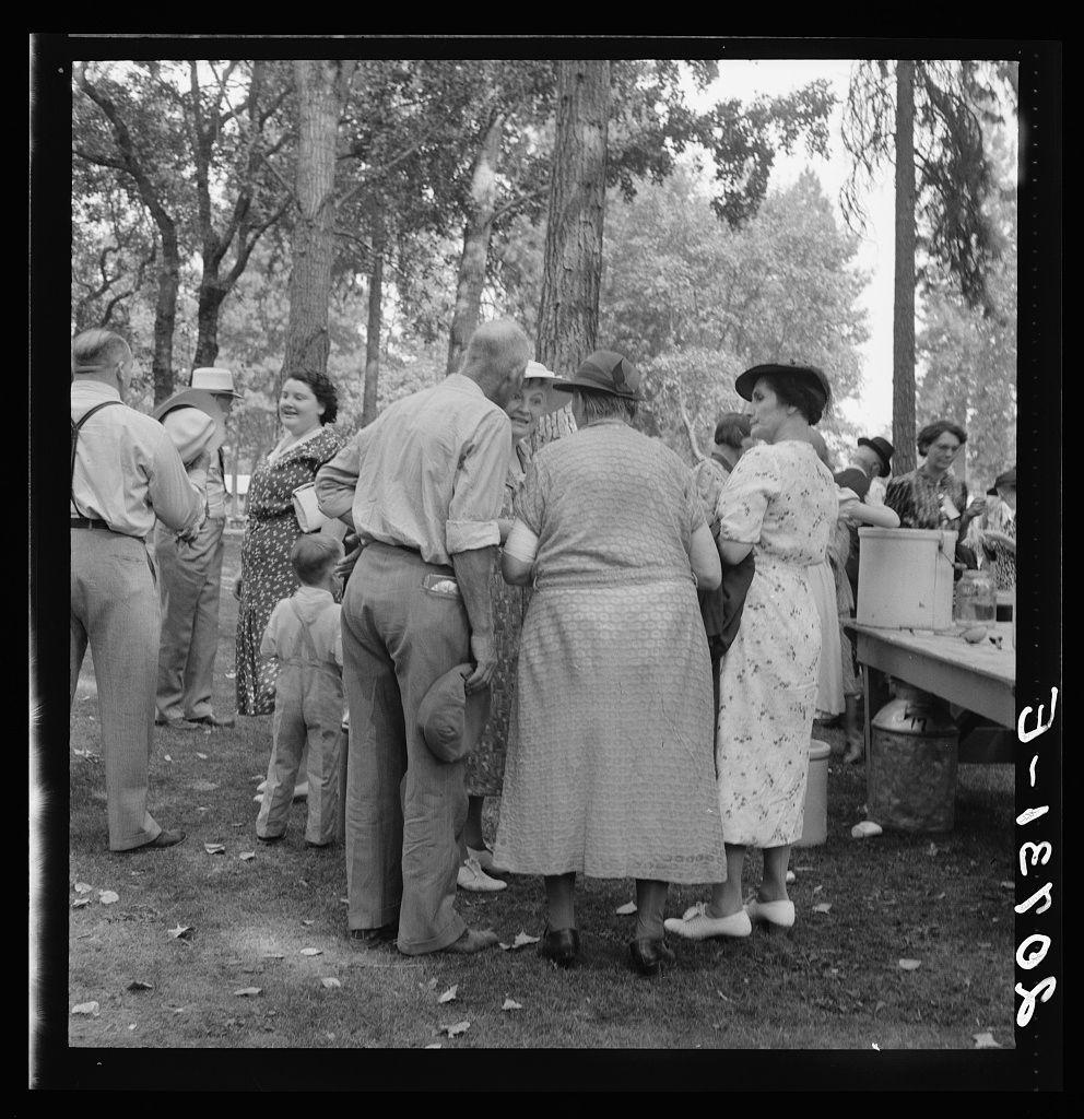 The image depicts a group of people gathered outdoors, possibly during an event or social gathering. The setting appears to be in a wooded area with trees providing shade for the attendees.

In the foreground, there is a woman wearing glasses and holding what seems to be a booklet or pamphlet. She has curly hair and wears dark-colored attire. To her left stands another woman dressed in a long coat over a patterned dress, interacting with someone out of frame on the right side. On the opposite end, near an outdoor serving table laden with food items like pitchers and possibly baked goods, there is a person wearing a hat.

In between these two women are other individuals: men standing casually or engaged in conversation; one holding what appears to be a drink cup. A child stands nearby, looking on without interacting directly with the others. The overall mood of the group seems relaxed and social.

The image conveys an intimate community atmosphere as people come together for this event, possibly after listening to speeches given that such gatherings were common post-speech events in public parks or similar venues during that period.