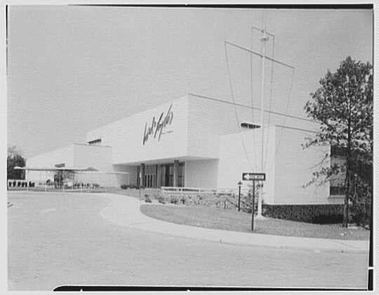 The image depicts a large, modern-style building with the name "Wanamaker's" displayed prominently on its facade. The architecture is characterized by clean lines and minimalistic design elements. In front of the building, there are some trees and shrubbery lining what appears to be an entrance road or drive leading up to it. To the right side of the photograph, a street sign can also be seen but the text is not legible in this view.

The photo appears black and white with soft shadows indicating that it was taken on a partly cloudy day. Given the presence of Gottschol-Schleisner Inc.'s watermark, it suggests that this image may have been used for professional purposes or documentation related to commercial architecture during its time. The date mentioned as June 7th in Lord & Taylor's business in Bala-Cynwyd could indicate a connection between this building and the department store chain Wanamaker's which operated locations across various cities, including Philadelphia where Balta Cynwyd is located.

Overall, it provides an historical snapshot of commercial architecture from mid-20th century America.