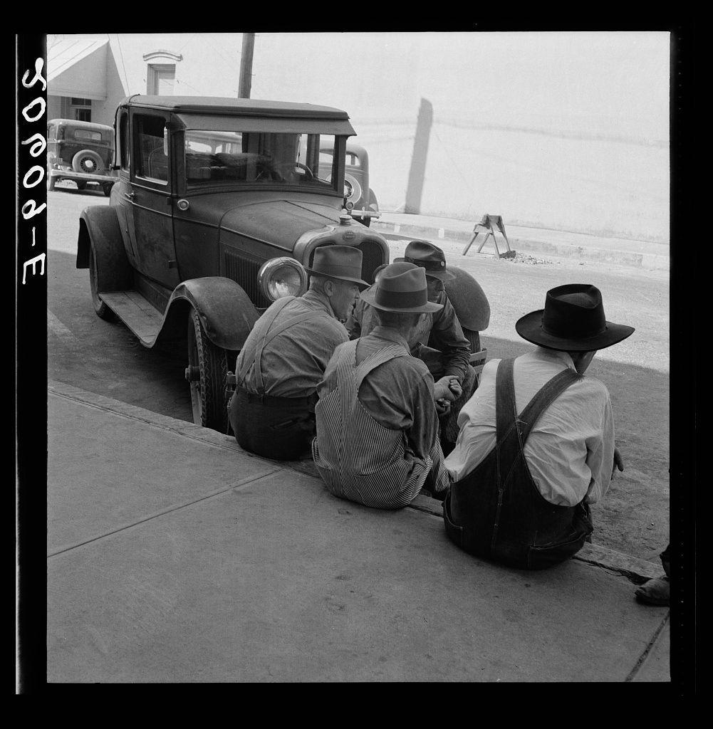 The image depicts a group of individuals sitting by the side of an older model car, engaged in conversation. The setting appears to be outside a building with a flat roof and large windows. Some people are wearing hats or caps while others have on suspenders over their shirts. A tripod is visible near one person's feet, suggesting that they may have been filming or photographing something earlier.

In the background, another vintage car can be seen parked further down the street. The image has a monochromatic color scheme typical of historical photographs from mid-20th century America and carries some markings in the top left corner which are not clearly legible but seem to indicate copyright information or identification numbers for archival purposes.

The photograph's composition, with its candid portrayal of everyday life and focus on ordinary people engaged in a social activity, is characteristic of Dorothea Lange's work. Her photographs often captured scenes from rural America during the Great Depression, focusing on themes such as poverty, labor, migration, race relations, and war."

In summary, this photograph showcases a moment among farmers or workers gathered around their car at what seems to be a community meeting in Independence, Oregon, which could provide insights into social dynamics, working conditions, and rural life during the early part of the 20th century.