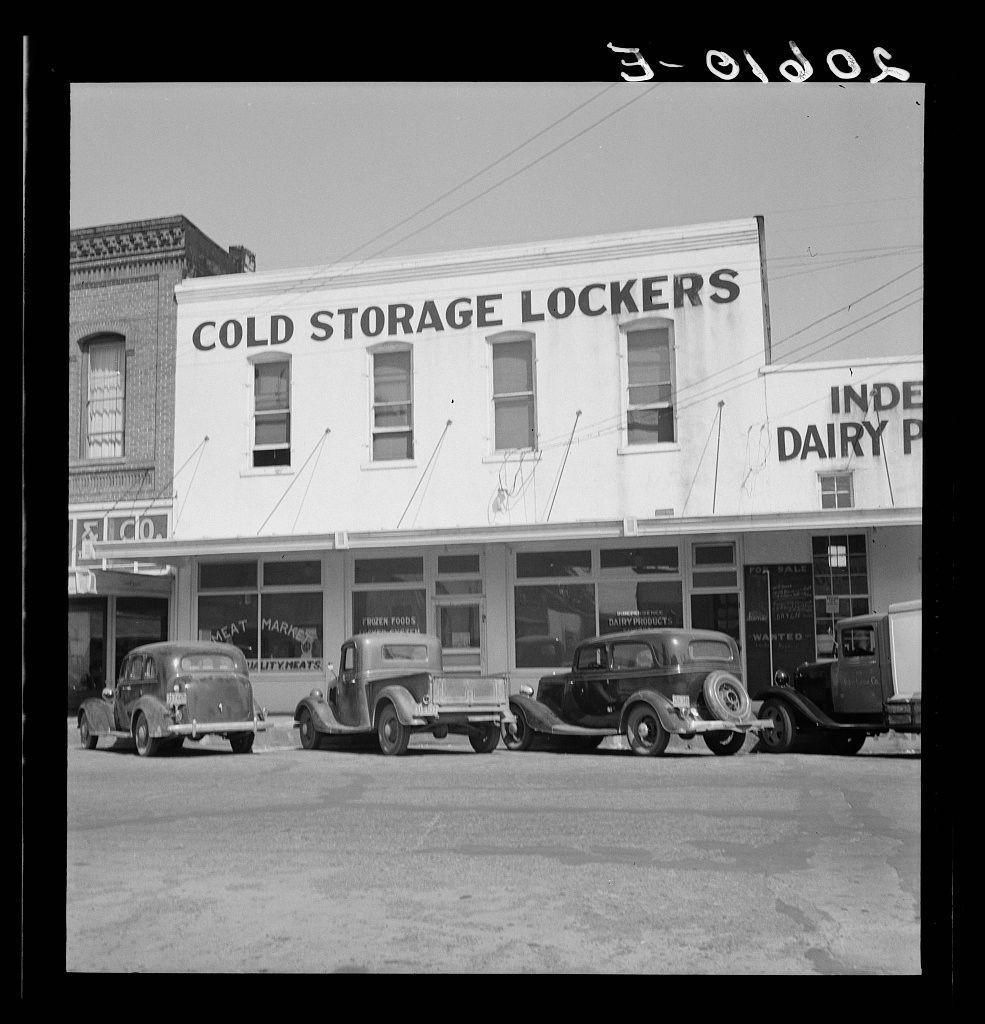 The image depicts a street scene from what appears to be the early 20th century. In front of a large, white building with windows and an awning, there are four vintage vehicles parked in two rows: three sedans on one side and one delivery truck or van on the other. The sign above the entrance reads "COLD STORAGE LOCKERS," indicating that this establishment provided refrigerated storage for perishable goods such as meat and vegetables to farmers who could then eliminate much home canning and preserving of foods, according to historical context mentioned in a caption.

The architecture of the building suggests it might have been an important commercial hub at the time. The clear sky indicates fair weather conditions during this period. There are no visible people or significant activity around the vehicles or on the street itself. Notably, the image is presented as black and white photography which adds to its historical feel.

In summary, this picture represents a piece of American history showcasing rural commerce from an earlier era with a focus on storage facilities for agricultural goods in small towns like Independence, Oregon.