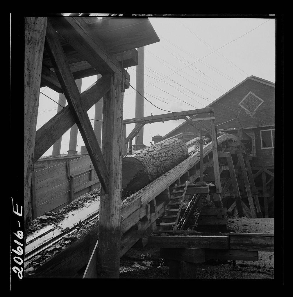 The image displays a black and white photograph of an industrial timber processing site. A large log is being transported on a series of wooden beams, part of what appears to be a chute or conveyor system used for moving logs from one location within the mill complex to another. The structure consists of several levels with multiple sets of tracks running parallel to each other at different heights. There's visible wear and tear on both the machinery and supporting structures.

In the background, there is part of an industrial building with a pitched roof covered in snow or frost, suggesting cold weather conditions. The sky above seems overcast which further indicates cooler temperatures. Electrical lines run horizontally across the upper portion of the image against a grayish backdrop that could be indicative of either cloudy skies or atmospheric mist.

The photograph carries a sense of historical significance and hard labor inherent to early timber processing industries. This particular setting is described as being part of the Pelican Bay Lumber Company mill, located near Klamath Falls in Oregon according to provided additional information from an external source (https://images.loener.nl/DorotheaLange/full/6495/6495b7baba25398d90c4f160.jpg).